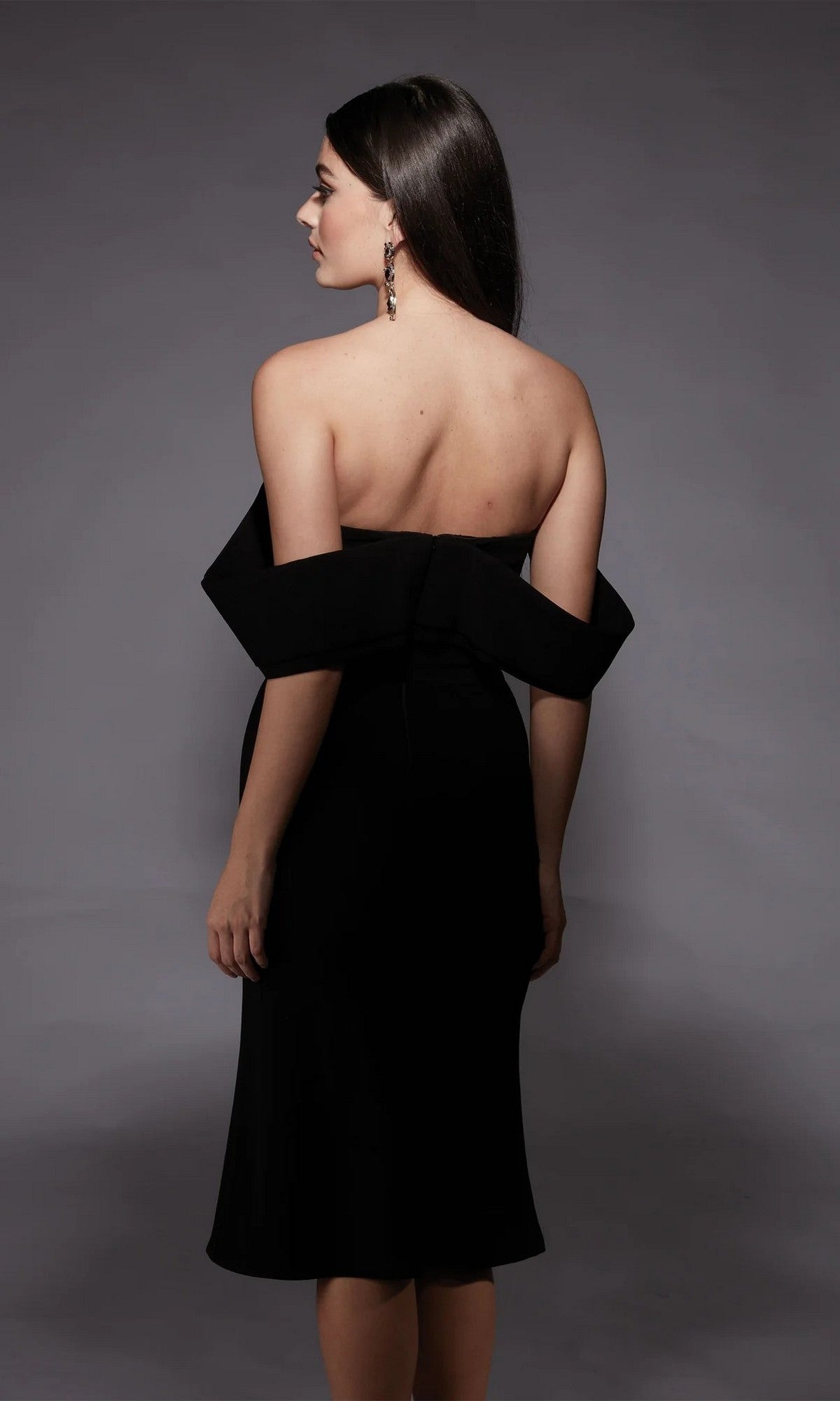 A woman with long dark hair, wearing the Alyce 70055 knee-length semi-formal dress and earrings, stands facing away against a plain gray background.